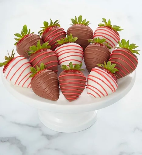 A white bowl holds a dozen strawberries that are dipped in either black or white chocolate and have a red swirl