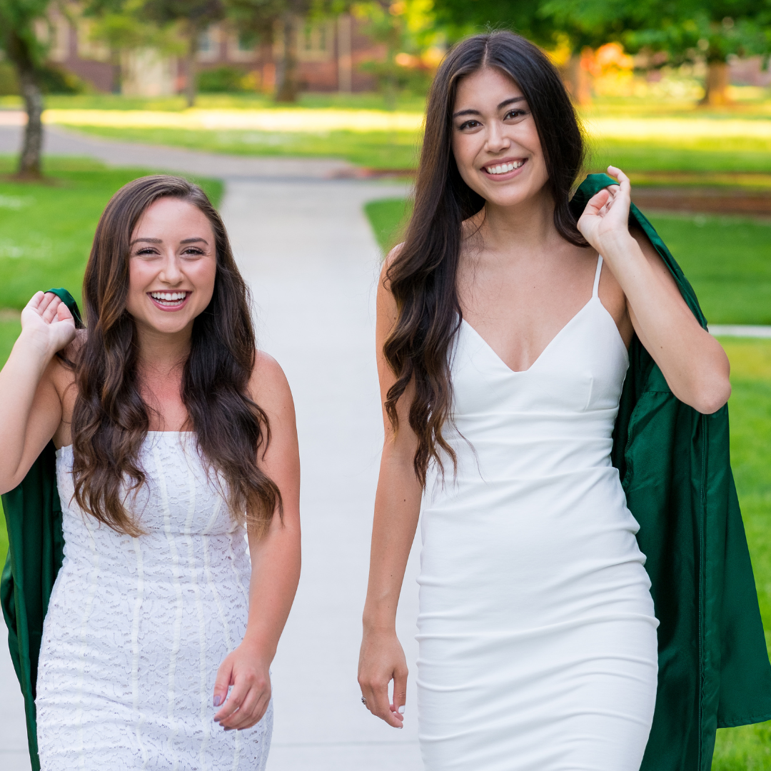 Two girls in white dresses and smiling holding their graduation gown over their shoulder